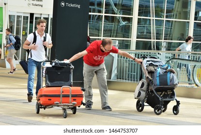 Denver, CO, USA. July 27, 2019. Young Man At Denver International Airport Struggling To Keep Control Of His Luggage Cart And Baby Buggy At The Same Time. 