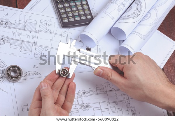 Department of engineering design.  Man\'s hand with the\
bearing. Mechanical engineer at work. Workplace of engineer.\
Drawings, pencil, pen, protractor, compass bearings, calculator on\
the table. 