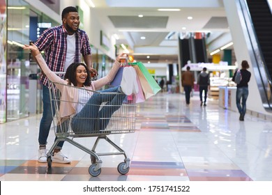 Department Store Fun. Happy african couple enjoying shopping together, man pushing shopping cart with girlfriend inside, fooling in mall, free space