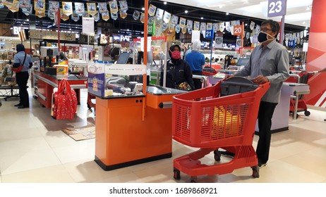 Depok, West Java, Indonesia - March 27th, 2020: A Male Customer With A Face Mask Stands In Front Of The Supermarket Cashier Staff With His Groceries On A Red Trolley