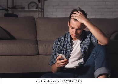 Deppresed teen guy checking mobile phone sitting on the floor in dark room, copy space