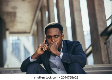 Depressed African American Businessman Telling Sad News Talking On Cell Phone Sitting On Office Stairs In Business Suit