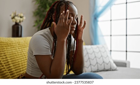 Depressed african american woman with braids sitting alone on sofa at home, unhappy expression reveals headache ache and stress - Shutterstock ID 2377535837