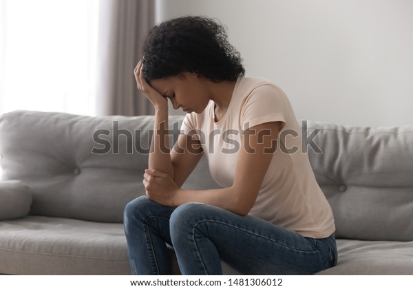 Depressed african American young woman sit on couch at home feel desperate heartbroken suffering from divorce, stressed upset black female thinking having life problems, miscarriage abortion concept