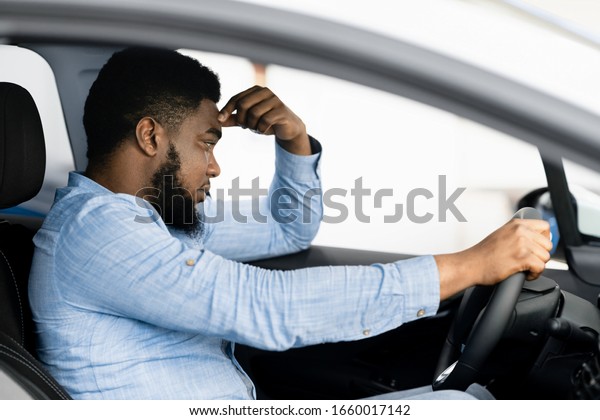 Depressed African Man Sitting In Driver\'s Seat Of\
New Too Expensive Car, Thinking Of Money Problems In Auto\
Dealership Store