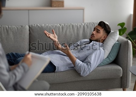 Depressed arab man lying on couch at psychologist's office, having session with counselor, millennial guy suffering from mental disorder, seeking professional help
