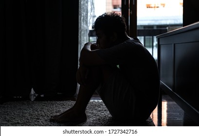 Depressed Asian man sitting on the floor hugging knees and looking away distraught with sadness. Loneliness sad man living alone and listening music from headphone. Mental health and drug abuse.