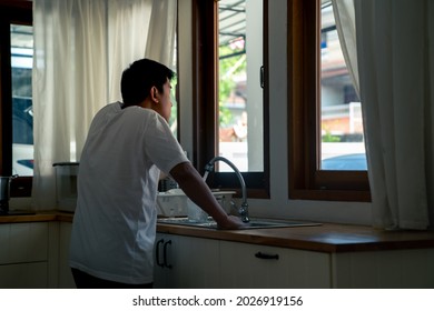 Depressed Asian Man Standing By The Window In The Kitchen With Stressed And Hopeless Face. Loneliness Sad Man Living Alone At Home With Negative Emotion. Mental Health And Life Problem Concept