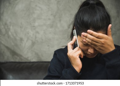 Depressed Asian Woman Sitting In The Dark Room Hand In Head And Talking On Phone Feeling Depressed, Lonely, Dramatic And Sad.