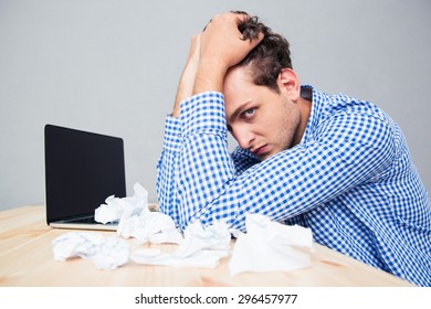 Depressed businessman sitting at the table with laptop and crumpled papers over gray background - Shutterstock ID 296457977