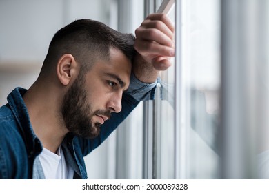 Depressed Caucasian Man Losing Job And Heartbroken At Same Time Alone, Feeling Bad And Expresses Negative. Unhappy Attractive Breaded Young Male Looks Outside Window In Living Room Interior, Close Up