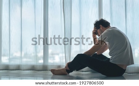 Depressed caucasian man losing job and heartbroken at same time sitting alone near window in dark at evening time with low light environment, PTSD Mental health and depression concept.