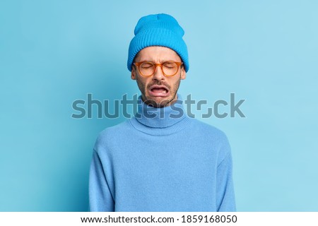 Depressed crying man being in stressful situation stands desperate as faces problem feels regret blames himself in failure wears trendy blue hat turtleneck spectacles poses indoor. Pessimistic person Foto stock © 