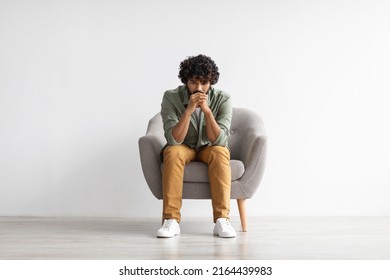 Depressed curly bearded young indian guy in casual outfit sitting in armchair over white studio background, looking down, thinking about something, suffering from loneliness, copy space, full length - Shutterstock ID 2164439983