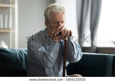 Depressed disabled retired elderly 50s grandfather hold in hands walking stick sit alone on couch looking lonely and unhappy, health problems, physically handicapped person, movement disorder concept