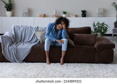 Depressed exhausted young African woman feeling stress, suffering from headache, sitting on cough at home, touching head, going through apathy, grief, loss. Depression, fatigue concept