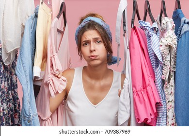 Depressed female standing near wardrobe rack full of clothes, having difficult choice not knowing what to put on. Nothing to wear concept. Choosing clothes for special occasion. Searching for clothing