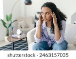 Depressed frustrated young brazilian or hispanic woman, sitting alone on a sofa, suffering from headache, migraine, feeling stressed, tired, thinks over bad news, problems in relationships