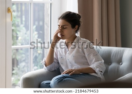 Depressed frustrated young Indian woman suffering from headache, migraine, touching face, head with closed eyes, feeling stressed, sick, tired, thinking over bad news, problems, crisis