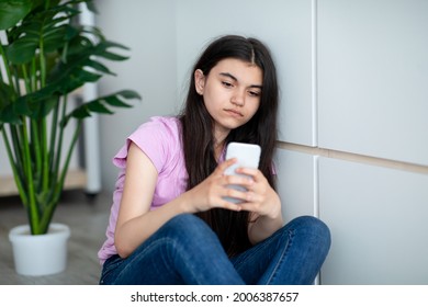 Depressed Indian Teen Girl With Smartphone Feeling Lonely, Looking At Screen With Sad Face Expression, Sitting Near Wall At Home. Upset Adolescent Suffering From Cyber Bullying Online