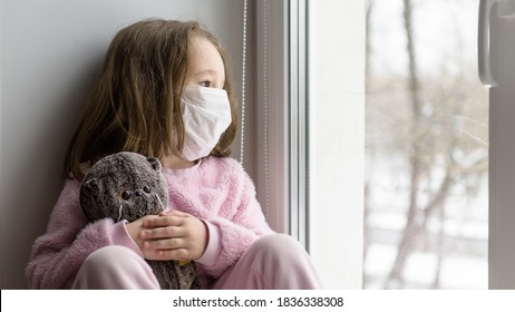 Depressed Kid In Medical Mask Looks Out Window, Sad Child Sits On Windowsill In Winter. Little Girl With Toy During Quarantine Safe From Corona Or Monkeypox Virus. Hospital, Pox And COVID-19 Concept.