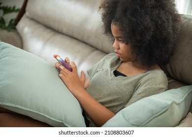 Depressed Little African Child Girl Sitting On Sofa In Living Room With Looking On Mobile Phone Screen With Sadness Face. Social Issue, Family Problem, Cyberbullying And Children Mental Health Concept