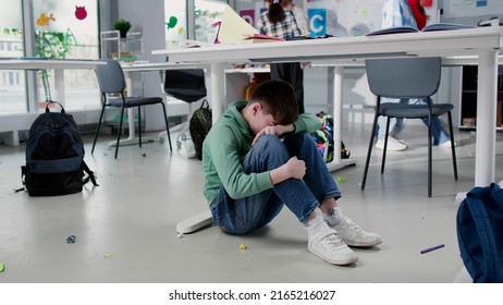 Depressed lonely boy sitting under desk in classroom with classmates playing on background having break - Shutterstock ID 2165216027