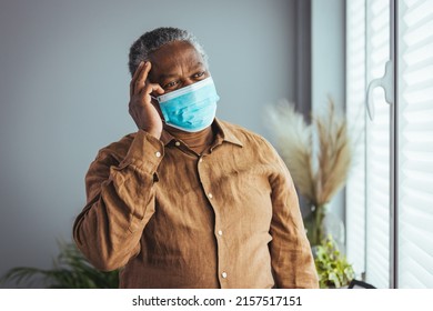 Depressed Looking Old African American Man Staying In Quarantine, Wearing A Protective Face Mask And Looking Through The Window Of His Home, Waiting For The Symptoms Of Corona Virus To Pass.