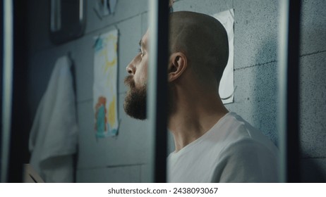 Depressed male prisoner looks at pictures of family or kids sitting on bed in prison cell. Illegally convicted man serves imprisonment term in jail. Detention center or correctional facility. Portrait