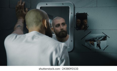 Depressed male prisoner stands in prison cell, looks at the pictures with family hanging near the mirror. Inmate serves imprisonment term for crime in jail. Detention center or correctional facility.