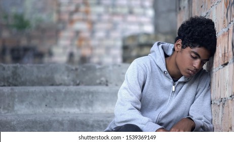 Depressed male teenager leaning brick wall sitting stairs, domestic violence