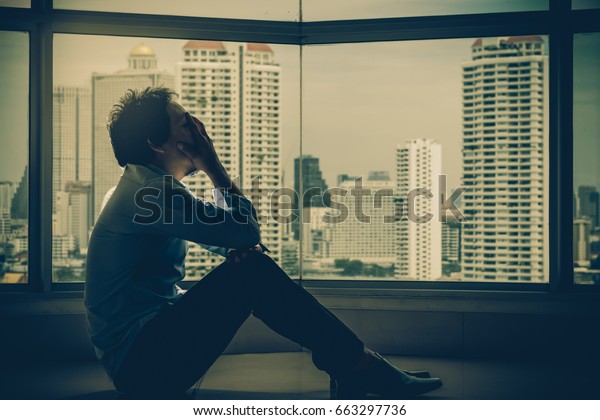 都市の風景の背景に窓の横にある低い光の環境で 落ち込んだ人が頭を抱えて室内の高層ビルに座り ドラマチックなコンセプト の写真素材 今すぐ編集