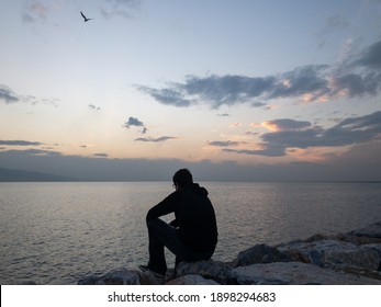 A Depressed Man Is Wearing Face Mask Due To Covid And Sitting Alone On The Bench By The Sea, Looking Across To The Sea Horizon. Missing Someone. Thinking About Someone.