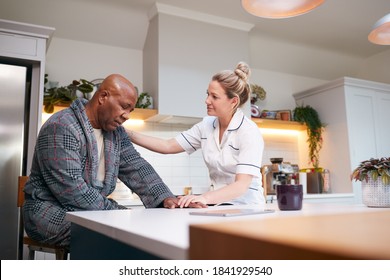 Depressed Mature Man In Dressing Gown Talking With Female Nurse In Kitchen At Home