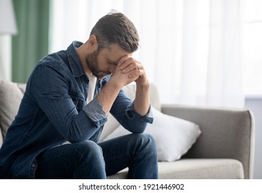 Depressed middle-aged man sitting on couch in living room, leaning on his hands, having financial troubles during quarantine or suffering from loneliness, copy space, home interior