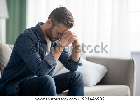 Depressed middle-aged man sitting on couch in living room, leaning on his hands, having financial troubles during quarantine or suffering from loneliness, copy space, home interior