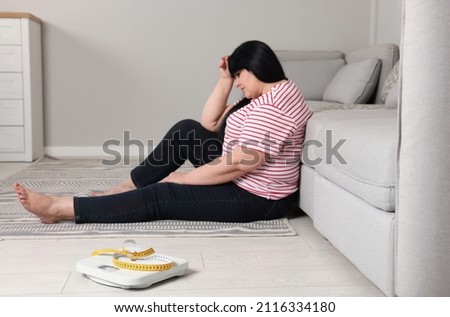 Depressed overweight woman sitting near sofa at home, focus on scales with measuring tape
