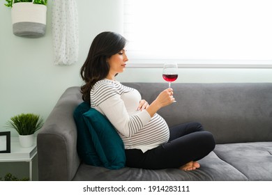 Depressed pregnant woman deciding whether or not to drink a glass of wine while sitting on the sofa. Caucasian expectant mother with bad and unhealthy habits