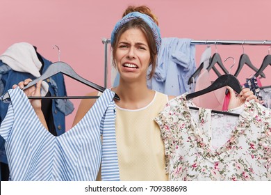 Depressed Sad Female Standing At Wardrobe Holding Two Hangers With Pieces Of Clothing Feeling Stressed As She Doesn't Know What To Wear On Job Interview, Looking At Camera With Mournful Expression