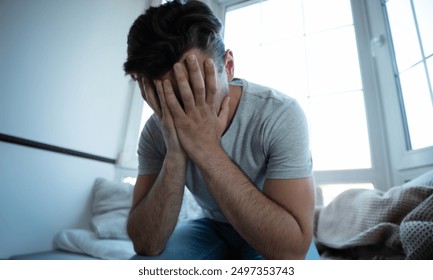 Depressed, sad, frustrated, crying, scared, instability mental and bipolar young man sitting on the windowsill at home. Headache because overworked and stress
