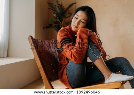 Depressed sad girl feeling lonely sitting by home winter looking out the window in winter. Asian woman feeling unstable in mental health. Concept of depression, loneliness, anxiety, grief emotions.