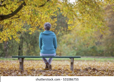 Depressed And Sad Old Woman On Bench In Autumn Park, 
From Back Angle