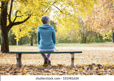 Depressed And Sad Old Woman On Bench In Autumn Park, From Back Angle