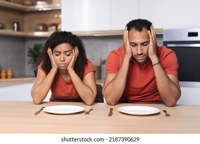 Depressed Sad Pensive Millennial African American Wife And Husband Hold Head With Hands Look At Empty Plates At Table In Kitchen. Diet, World Food Crisis, Waiting For Dinner And Hunger Family