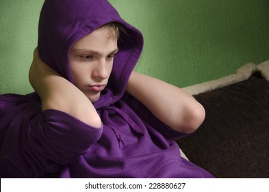 Depressed teenager with arms around his neck and hood on head, looking down.