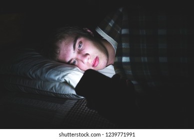 Depressed teenager browsing the internet on his mobile phone as he is lying on his bed in the dark.