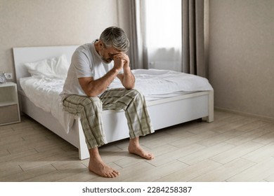 Depressed unhappy frustrated grey-haired mature man wearing pajama barefoot sitting on bed in the morning, leaning on his knee, suffering from mental breakdown, bedroom interior, copy space - Powered by Shutterstock