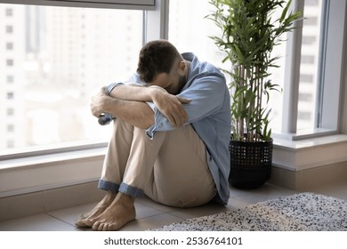 Depressed unhappy young man sitting at window on floor, bending head forward, hiding face, feeling desperate, frustrated about money loss, bankruptcy, divorce, grief, needing psychological support - Powered by Shutterstock