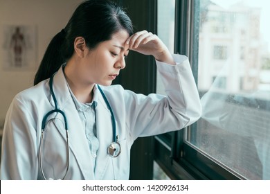 Depressed Woman Doctor Leaning Against Window Upset With Bad Surgery Result In Therapist Office. Frowning Sad Young Female Medical Staff Sad About Patient Death Fail Medicine Concept Clinic Hospital