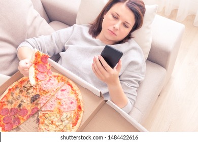 Depressed Woman Eating Pizza And Holding Phone Laying On Sofa At Home, Warm Yellow Tone